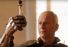 Swen Vincke in a full suit of armour, holding up a potion to the light.