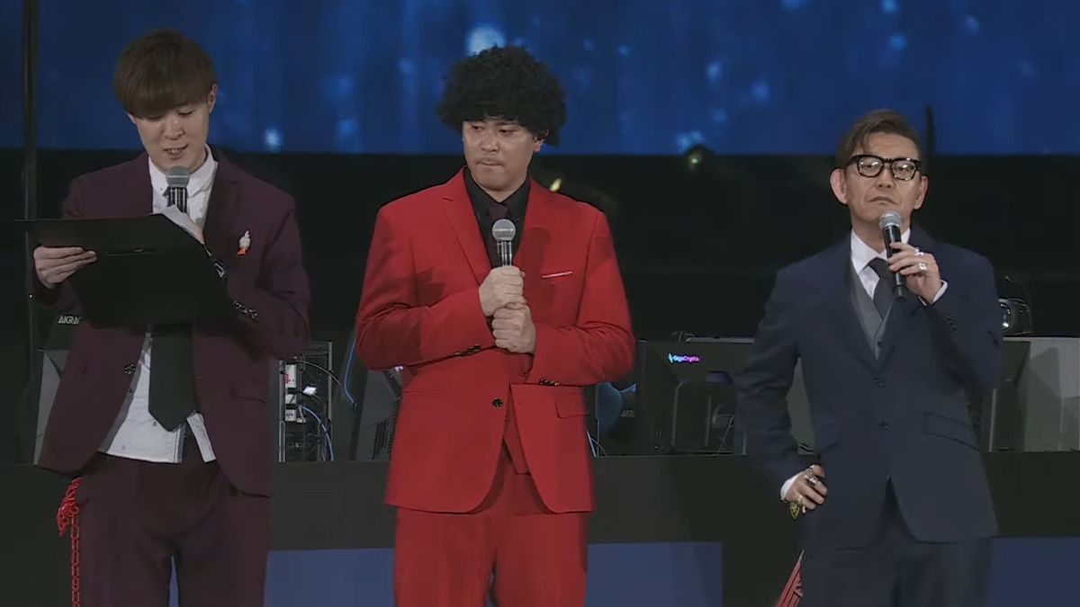 Naoki Yoshida stands on the right with his hand on his hip, wearing a blue suit.,